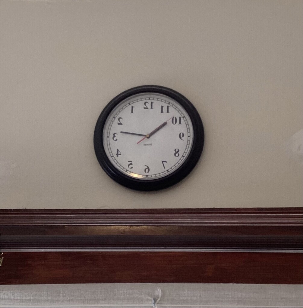 A mirror-reversed clock hanging on the wall. The numbers run counterclockwise and they are drawn backwards.