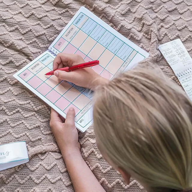 a kid writing with a pen in a colorful ledger book with columns labelled "date" "description" "transaction"