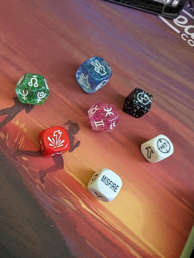 A photo of seven dice that have been rolled. One depicts a hand holding two fingers up, two depict astrological(?) symbols, one depicts a sort of zap symbol with lines radiating from the point of impact, one says "MISFIRE", one says "HIT" in a crosshairs symbol, and one has a sort of swirly symbol (and other faces on it are clearly blank).