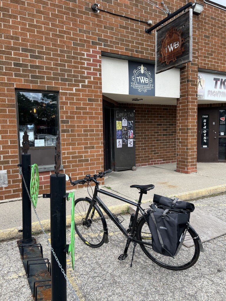 In the foreground a bike is upright on its kickstand. It has panniers over the rear wheel. In the background, a sign hangs in front of a red brick wall over a building entrance. It reads “TWB Cooperative Brewing”