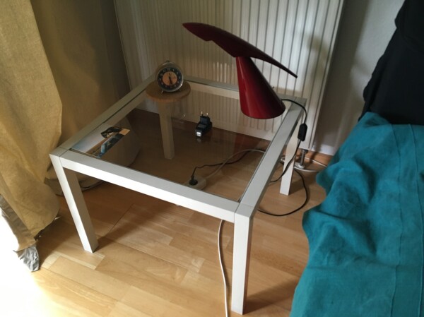 square low IKEA table with a glass plate, there’s a red lamp and an unused alarm clock on it (and an USB charger)