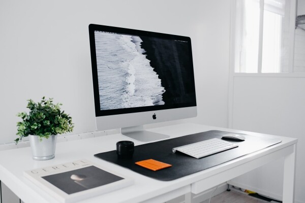minimalist desktop in all white bright room on a white desk, there an Apple computer, wireless keyboard and mouse on a big pad, and a plant and book next to that