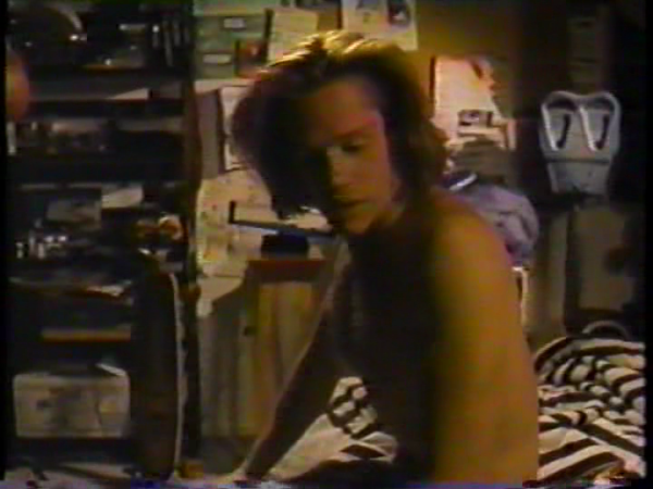 A lower resolution screen capture of a shirtless guy with Bon Jovi hair waking up.  There's 90s stuff everywhere, including a stolen parking meter.