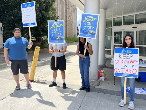 Four people with picket signs standing looking at the camera. The signed say things like “LCBO Workers On Strike” and “Keep Our Money In Ontario”